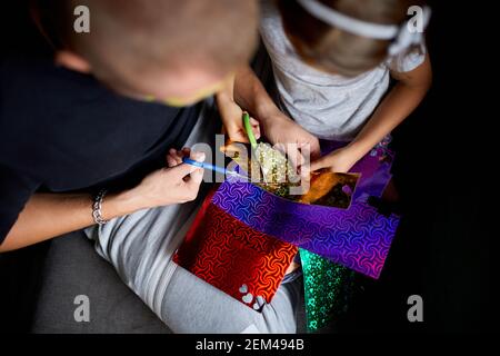 Daughter and father having fun making craft together at home on the sofa, cutting a paper with scissors, dark light, Fathers day, family. Stock Photo