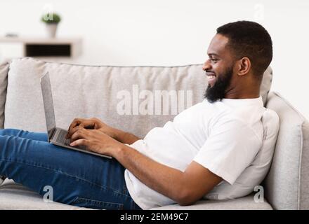 Black Freelancer Guy Working On Laptop Lying On Sofa Indoor Stock Photo