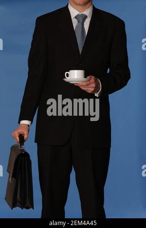 Faceless white business man in a black suit with a blue tie carrying a black leather briefcase and tea cup against a blue background Stock Photo