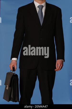Faceless white business man in a black suit with a blue tie carrying a black leather briefcase against a blue background Stock Photo