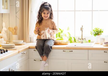 Adorable Little Arab Girl Eating Cookies And Drinking Milk In Kitchen Stock Photo