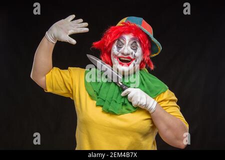 Scary clown with a horrible make-up laughs and with big knives in hands on a black background Stock Photo