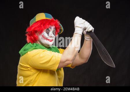 Scary clown with a horrible make-up laughs and with big knives in hands on a black background Stock Photo