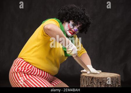 Scary clown with a horrible make-up laughs and with big knives in hands on a black background Stock Photo