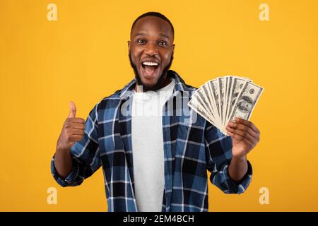 Joyful Black Guy Holding Money Cash Gesturing Thumbs-Up, Yellow Background Stock Photo