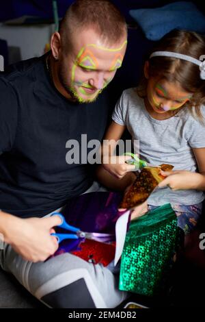 Daughter and father having fun making craft together at home on the sofa, cutting a paper with scissors, dark light, Fathers day, family. Stock Photo