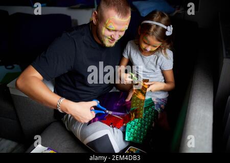 Daughter and father having fun making craft together at home on the sofa, cutting a paper with scissors, dark light, Fathers day, family. Stock Photo