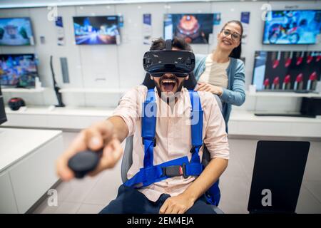 Young cute playful amazed couple having fun with VR goggles in the tech store. Stock Photo