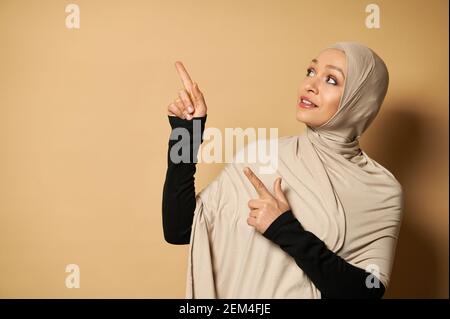Beautiful Muslim woman in hijab pointing her index fingers up to the side of beige background with copy space Stock Photo
