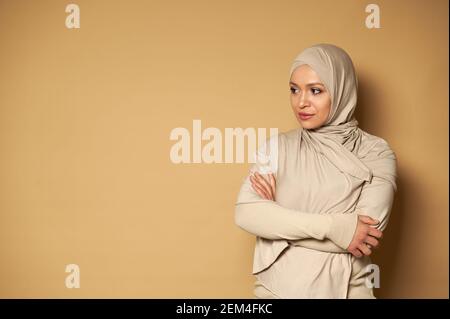 Portrait of serene beautiful Muslim woman in hijab and beige traditional formal dress looking to the side and crossing her arms in front of her on bei Stock Photo