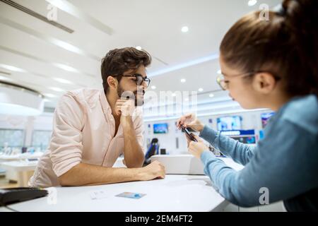 Close up view of stylish bearded hipster curious man getting instructions for a mobile from professional girl seller in a tech store.