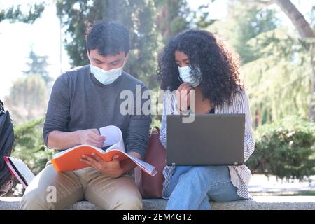 Two latin students wearing protective face mask studying together sitting on a bench outdoor. New normal at university campus. Stock Photo