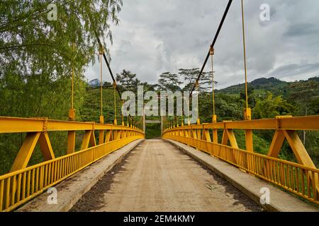 Old suspension bridge with yellow paint and a single track leading into the dense forest of Jardin in Colombia, South America Stock Photo