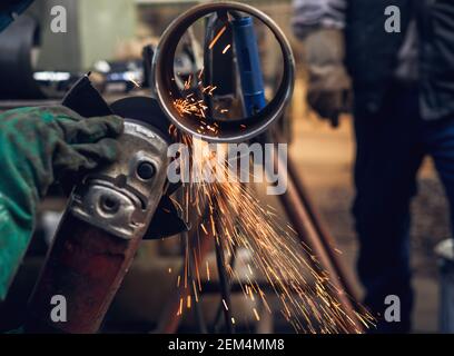 Side close up view of professional hardworking man hands cuts metal pipe with a large electric grinder while sparks flying in the industrial fabric wo Stock Photo