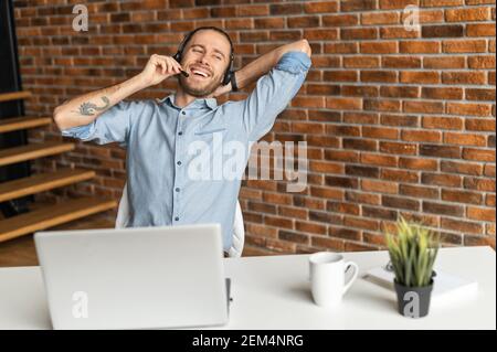 Smiling man with headset sitting in front of desk with a laptop and talking on his break, has a tattoo, talking with friends, colleague, dressed in a casual style, has a pleasant conversation, at home Stock Photo
