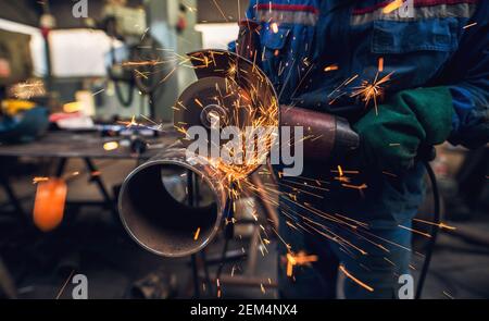 Side close up view of professional hardworking man in uniform cuts metal pipe sculpture with a large electric grinder while sparks flying in the indus Stock Photo