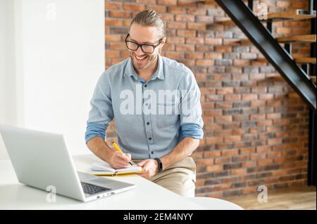 Young diligent employee with glasses sitting at the table and writing down in the notebook tasks put on, looking at the laptop screen, working online in the loft style apartment, consulting client Stock Photo