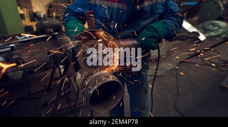Side close up view of professional focused hardworking man in uniform cuts metal tube sculpture with a large electric grinder while sparks flying in t Stock Photo