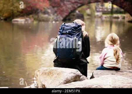 Rear view of a girl and a woman sitting on rocks along a river, New York City, New York State, USA Stock Photo