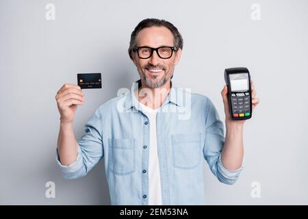 Portrait of handsome smart cheerful mature man holding in hands pos device bank card isolated over grey pastel color background Stock Photo