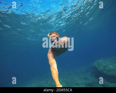 Underwater photo of a young sportive active man diving in the turquoise exotic sea near the rocks and taking a selfie with a stick for summer holidays Stock Photo