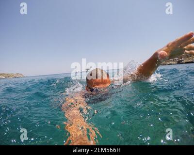 Young joyful man diver swimming in the aquatic exotic sea with goggles near seaside and yachts and in summer holidays. Stock Photo