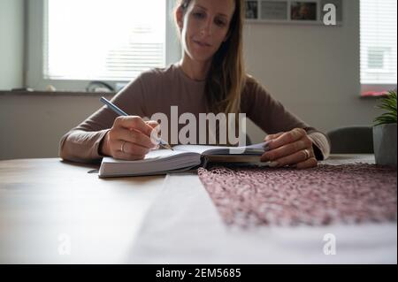 Young woman writing in notebook with a pencil sitting at home dining table. Stock Photo