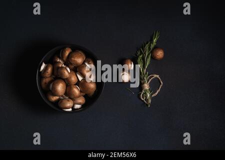 Bowl of raw baby bella mushrooms and bunch of fresh rosemary on dark grey background viewed from above, plant based food
