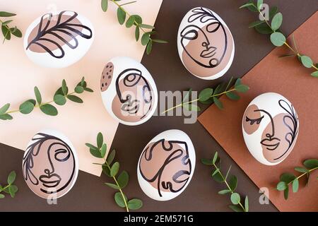 Happy Easter concept. Surreal faces on eggs and green branches of eucalyptus on brown background. Art und Online style. Top view Flat lay Copy space. Stock Photo