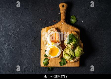 Poached egg and avocado on sliced toasted bread. Toasts on rustic wooden cutting board on dark grey background, top view Stock Photo