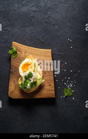 Poached egg and avocado on sliced toasted bread. Toast on rustic wooden cutting board on dark grey background, top view Stock Photo