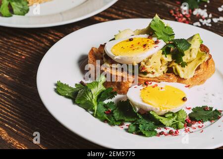 Poached egg and avocado  with fresh cilantro on sliced toasted bread. Toast on white dinner plate,  rustic wooden background Stock Photo