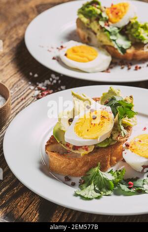 Poached egg and avocado  with fresh cilantro on sliced toasted bread. Toasts on white dinner plates, brunch for two people, rustic wooden background Stock Photo