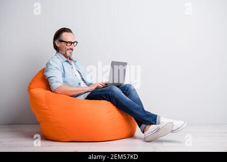 Profile side view of handsome clever cheerful mature man sitting on bean chair using laptop isolated over grey pastel color background Stock Photo
