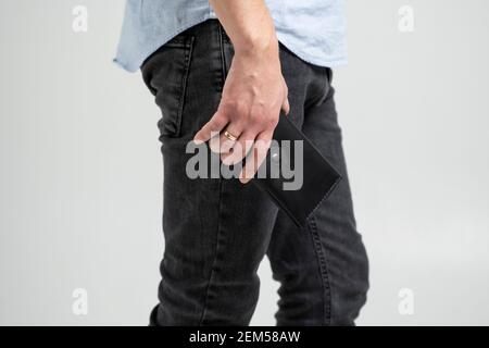 Stylish handmade black leather wallet in a mens hand in blue shirt and black jeans on white background. Stock Photo