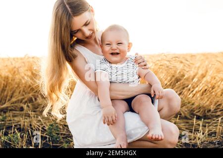 Happy beautiful mother with her baby on the golden wheat field, family on the grain field, mom hugs her little boy Stock Photo