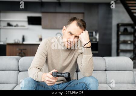 Upset young adult man sitting with joystick in hand, playing video games, game over, annoyed, on self isolation, at home, relaxing, hard day, long time loading, lost connection, no one to play with Stock Photo