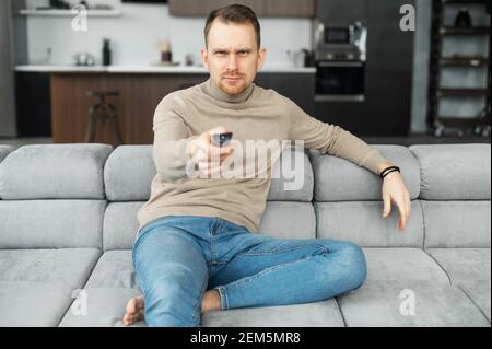 Young adult man is sitting on sofa with a TV remote in the hand, trying to watch something on TV, saw himself in the program, spending time at home, tuning the TV, fast forward or rewind, foreground Stock Photo