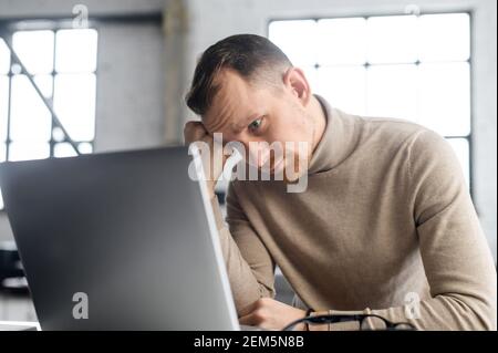 Young disappointed businessman sitting at the laptop, propped his head on his fist and watching at the computer screen, thinking about budget and office renovation, overworked, in a hopeless position Stock Photo
