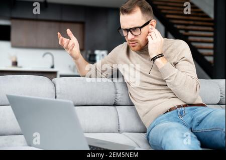 Disoriented young man looking at computer screen, holding the hand on the earphone, wrinkling the forehead, checking the connection, doesn't hear the companion, computer is broken, what happened Stock Photo