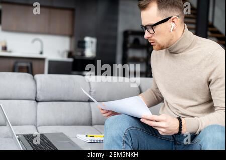 Serious businessman reviewing documents, schedules and reports, sitting on the kitchen, laptop on the sofa, work from home, in comfort clothes, analyze statistic, carefully examines every detail Stock Photo