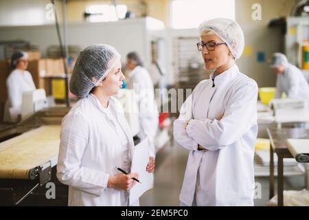Two dedicated female workers are discussing their daily quota while standing  near bakery production line. Stock Photo