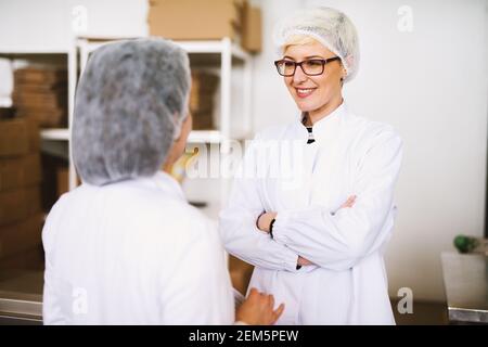 Two young joyful female workers in sterile cloths are having a chat while standing in factory cargo room. Stock Photo