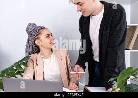 Young Caucasian business colleagues man and woman communicating in the office. Attractive woman with kanekalon pigtails sits at table with man standin Stock Photo