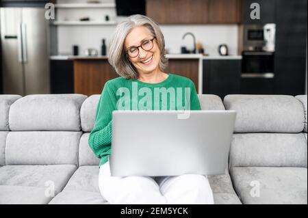 Smiling charming elderly woman looks at the screen of the laptop and laughs sitting on a couch at home, an elderly lady wearing eyeglasses spends leisure in networks, watching movies, has a video call Stock Photo