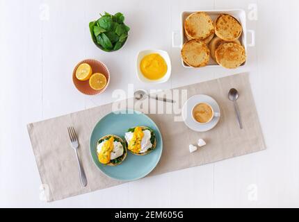 Eggs Florentine, English muffins, spinach, poached eggs, Mornay sauce, chive herbs, lemon, cup of coffee. White wooden table with breakfast. Stock Photo