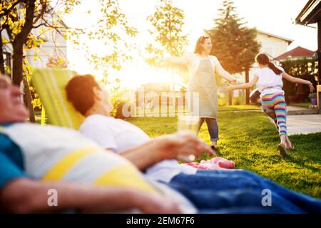 Cheerful woman is playing with her daughter in front of the house on a beautiful day while her parents are watching her. Stock Photo