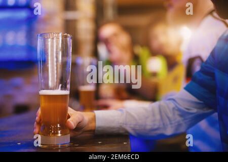 Half drank beer pint is being held by a soccer fan that sits at the bar in a local pub. Stock Photo