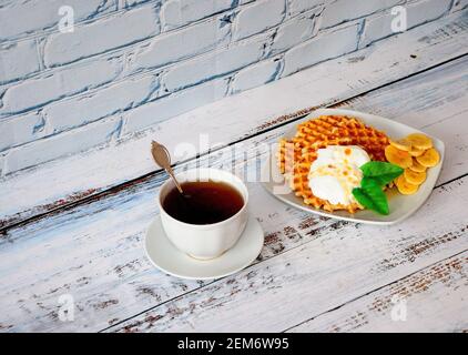 Sweet breakfast, a plate of waffles with protein cream and banana pieces and a cup of black tea on a wooden table. Close-up. Stock Photo