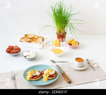 Eggs Benedict, English muffins, grilled ham, poached eggs, Hollandaise sauce, chive herbs, lemon, cup of coffee. White wooden table with breakfast. Lo Stock Photo